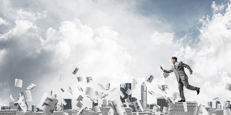 Businessman in suit running with phone in hand among flying paper documents with cloudly sky. Mixed media.の写真素材