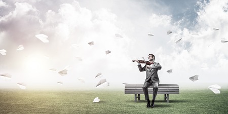 Young man wearing suit and glasses sitting on bench and playing violinの写真素材