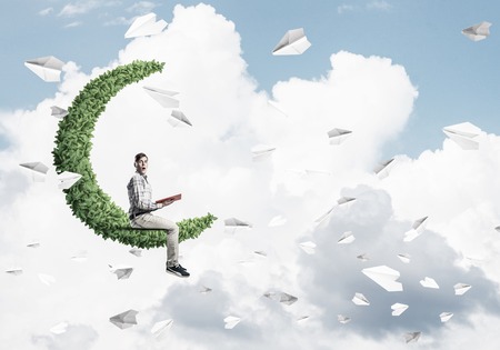 Young man in casual floating on green moon in blue sky with red book in handsの写真素材