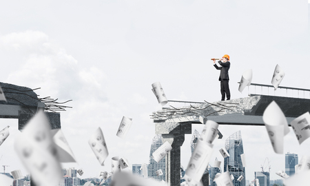 Young engineer in suit and helmet looking in spyglass while standing among flying papers on broken bridge with cityscape on background. 3D rendering.の写真素材
