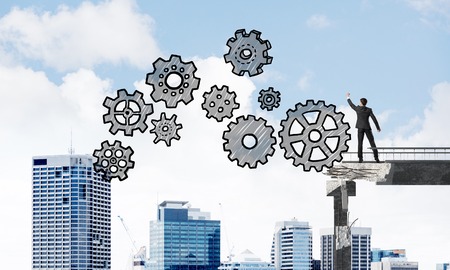 Businessman in suit drawing sketches of gear mechanism while standing on broken bridge with cityscape on background. 3D rendering.の写真素材
