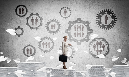 Business woman in suit standing on pile of documents among flying paper planes with social gear structure on background. Mixed media.の写真素材