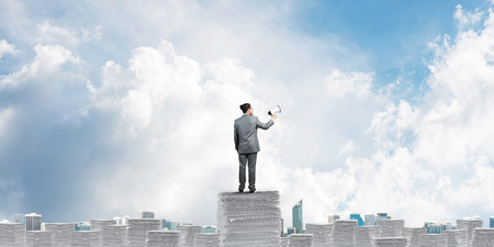 Businessman in suit standing on pile of documents with speaker in hand with skyscape and city view on background. Mixed media.の写真素材