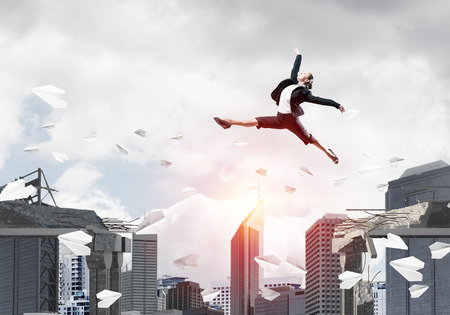 Business woman jumping over gap with flying paper planes in concrete bridge as symbol of overcoming challenges. Cityscape with sunlight on background. 3D rendering.の写真素材
