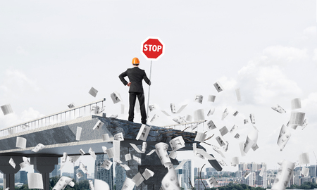 Rear view of engineer in helmet holding stop sign while standing among flying papers on broken bridge with cityscape on background. 3D rendering.の写真素材