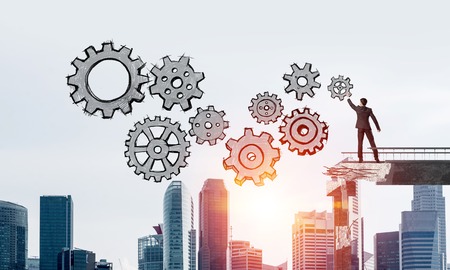 Businessman in suit drawing sketches of gear mechanism while standing on broken bridge with cityscape and sunlight on background. 3D rendering.の写真素材