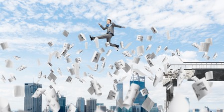 Businessman jumping over gap with flying paper documents in concrete bridge as symbol of overcoming...