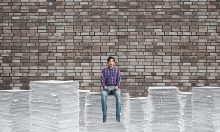 Young man in casual clothing sitting on pile of documents with brick wall on background. Mixed...