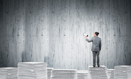 Businessman in suit standing on pile of documents with speaker in hand with grey wall on background. Mixed media.の写真素材
