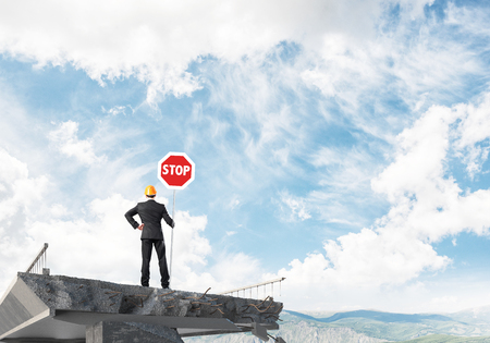 Rear view of engineer in helmet holding stop sign while standing on broken bridge with skyscape and nature view on background. 3D rendering.の写真素材