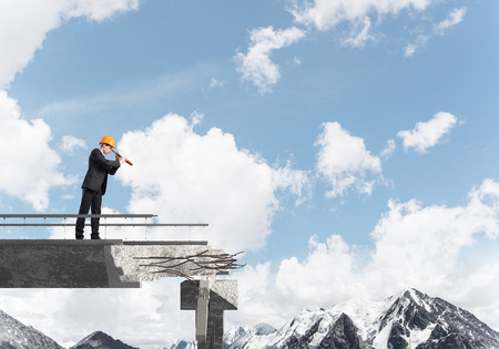 Young engineer in suit and helmet looking in spyglass while standing on broken bridge with skyscape and nature view on background. 3D rendering.の写真素材