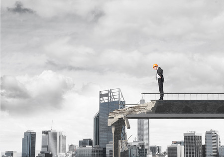 Young engineer in suit and helmet looking down while standing on broken bridge with cityscape on background. 3D rendering.の写真素材