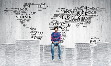 Young man in casual wear sitting on pile of documents with business-related terms in form of world map on background. Mixed media.の写真素材
