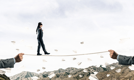 Businessman walking blindfolded on rope among flying paper planes and above high mountains as symbol of hidden threats and support. Nature view on background.の写真素材