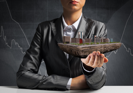 Cropped image of businessman in suit presenting in his hand modern city block with business sketches on background. 3D rendering.の写真素材