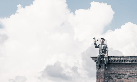 Young businessman sitting on roof and screaming emotionally in megaphoneの写真素材
