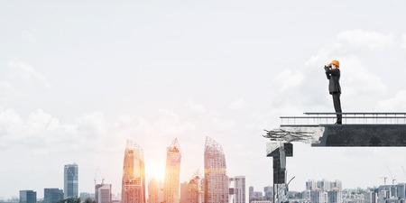 Engineer in suit and helmet looking in binoculars while standing on broken bridge with cityscape and sunlight on background. 3D rendering.の写真素材