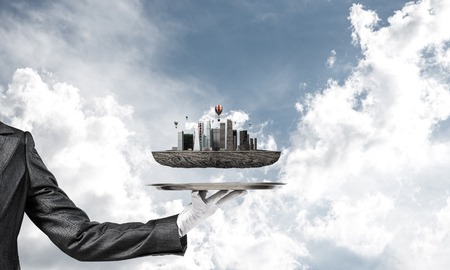 Cropped image of waitresss hand in white glove presenting modern city block on metal tray with blue cloudy skyscape on background. 3D rendering.の写真素材