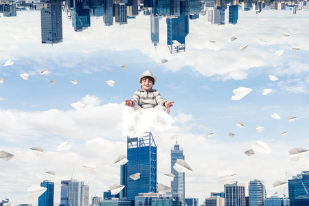 Young boy keeping eyes closed and looking concentrated while meditating on cloud among flying paper planes between two urban worlds.の写真素材