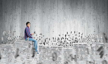 Young man in casual clothing sitting among flying letters with grey wall on background. Mixed media.の写真素材