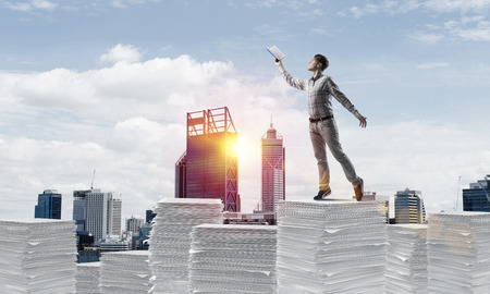 Man in casual wear keeping hand with book up while standing on pile of documents with cityscape and sunlight on background. Mixed media.の写真素材