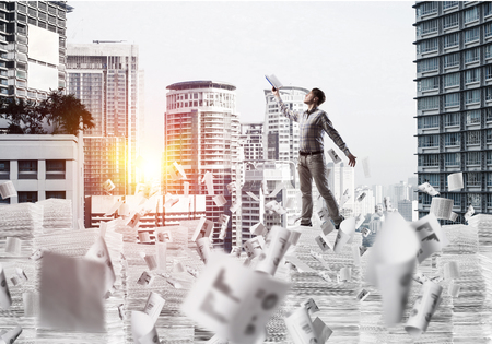 Side view of man in casual wear keeping hand with book up while standing among flying papers with cityscape and sunlight on background. Mixed media.の写真素材
