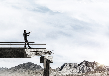 Businessman walking blindfolded on concrete bridge with huge gap as symbol of hidden threats and risks. Skyscape and nature view on background. 3D rendering.の写真素材