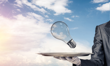 Cropped image of waitress's hand in white glove presenting lightbulb on metal tray as symbol of new idea with cloudy skyscape on background.の写真素材