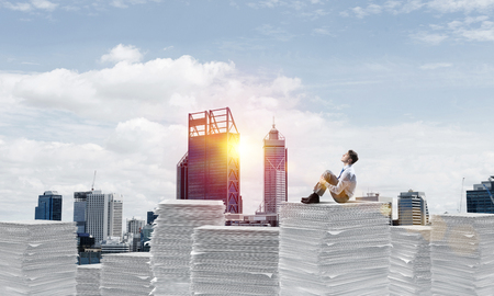 Thoughtful businessman sitting on pile of documents and looking away with cityscape and sunlight on background. Mixed media.の写真素材