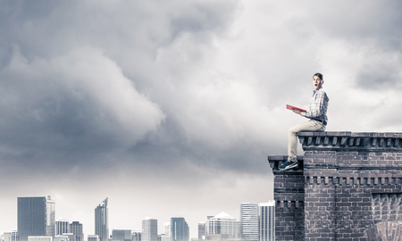 Young man in casual sitting on parapet with red book in handsの写真素材