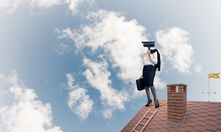 Faceless businesswoman with camera zoom instead of head standing on house roofの写真素材
