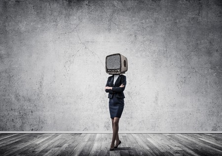 Business woman in suit with an old TV instead of head keeping arms crossed while standing inside empty room with gray wall on background.の写真素材