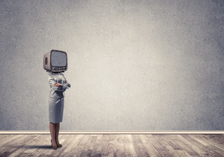 Business woman in suit with an old TV instead of head keeping arms crossed while standing inside empty room with gray wall on background.の写真素材