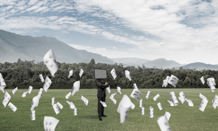 Businessman in suit with TV instead of head keeping arms crossed while standing on the road among flying papers with beautiful landscape on background.の写真素材
