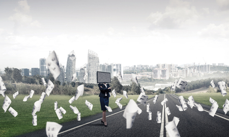 Business woman in suit with TV instead of head keeping arms crossed while standing on the road among flying papers with beautiful landscape on background.の写真素材