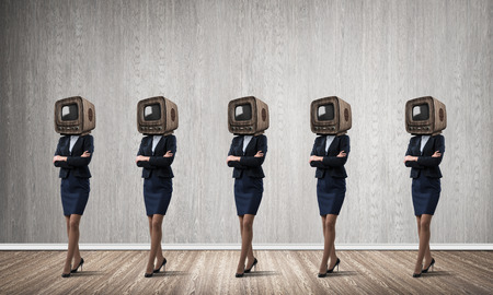 Business women in suits with old TV instead of their heads keeping arms crossed while standing in a row in empty room with gray wall on background.の写真素材
