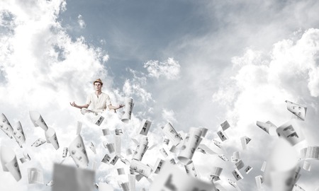Man in white clothing keeping eyes closed and looking concentrated while meditating among flying papers in the air with cloudy skyscape on background.の写真素材