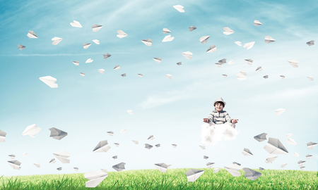 Young little boy keeping eyes closed and looking concentrated while meditating on cloud among flying paper planes with bright and beautiful landscape on background.の写真素材