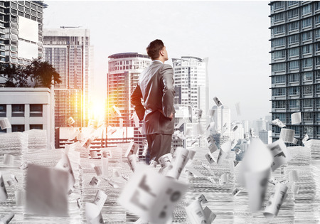 Attractive confident businessman in suit standing among flying letters with cityscape and sunlight on background. Mixed media.の写真素材