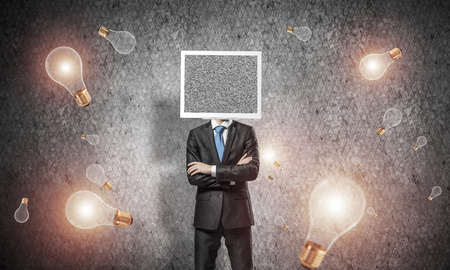 Businessman in suit with monitor instead of head keeping arms crossed while standing among flying lightbulbs and with gray dark wall on background.の写真素材