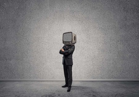 Businessman in suit with an old TV instead of head keeping arms crossed while standing inside empty room with gray wall on background.の写真素材
