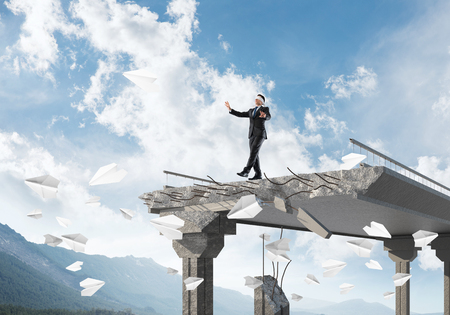 Businessman walking blindfolded among flying paper planes on concrete bridge with huge gap as symbol of hidden threats and risks. Skyscape and nature view on background. 3D rendering.の写真素材