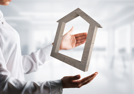 Business woman in white shirt keeping stone house symbol in hands with office view on background. Mixed media.の写真素材