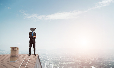 Faceless businessman with camera zoom instead of head standing on house roofの写真素材