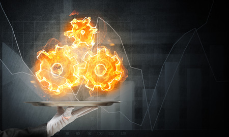 Cropped image of waitress's hand in white glove presenting flaming cogwheels structure on metal tray with dark wall on background.の写真素材