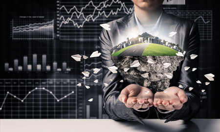 Businessman in suit keeping green island with skycraper city and flying paper planes in hands with business sketches on background.の写真素材