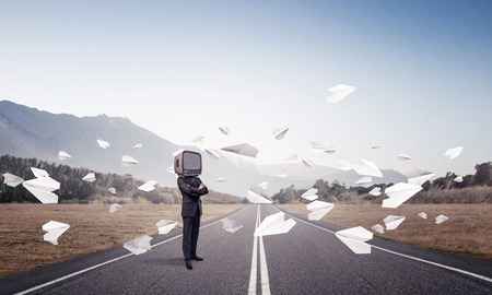 Businessman in suit with old TV instead of head keeping arms crossed while standing on the road among flying paper planes with beautiful landscape on background.の写真素材