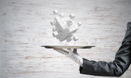 Cropped image of waitresss hand in white glove presenting multiple cubes on metal tray with gray wall on background. 3D rendering.の写真素材