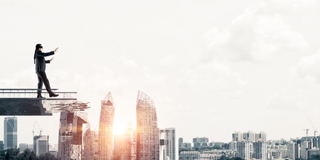 Businessman walking blindfolded on concrete bridge with huge gap as symbol of hidden threats and risks. Cityscape view with sunlight on background. 3D rendering.の写真素材