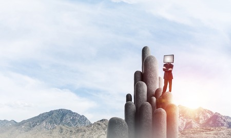 Businessman in suit with TV instead of head keeping arms crossed while standing on the top of stone columns with beautiful landscape on background.の写真素材